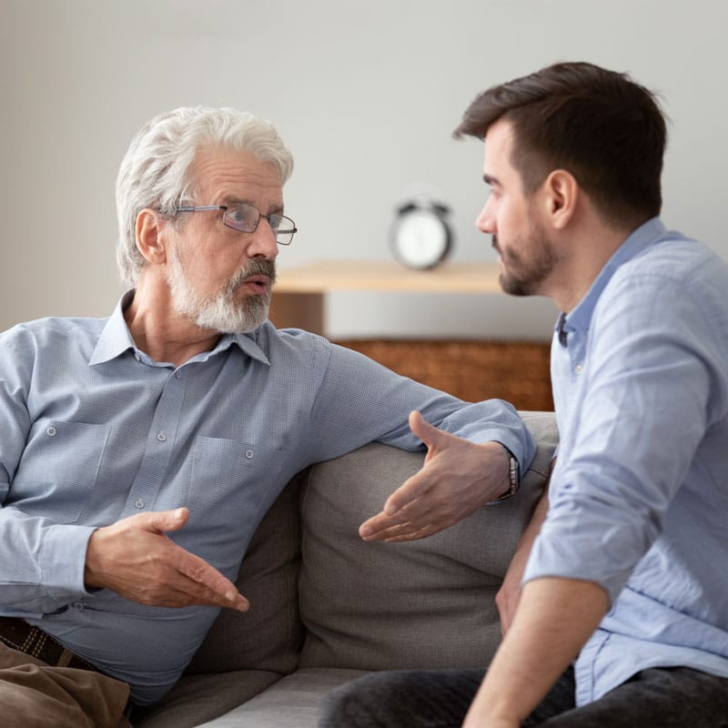 an elederly man speaking with his hands to a younger man on a couch inside of a Certified Community Behavioral Health Clinic (CCBHC)